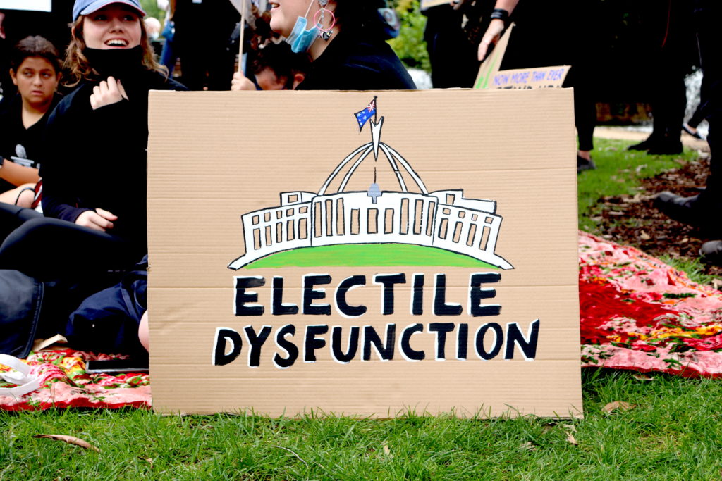 A cardboard sign from Naarm's March4Justice reads 'Electile Dysfunctino' under a painted image of parliament house. 