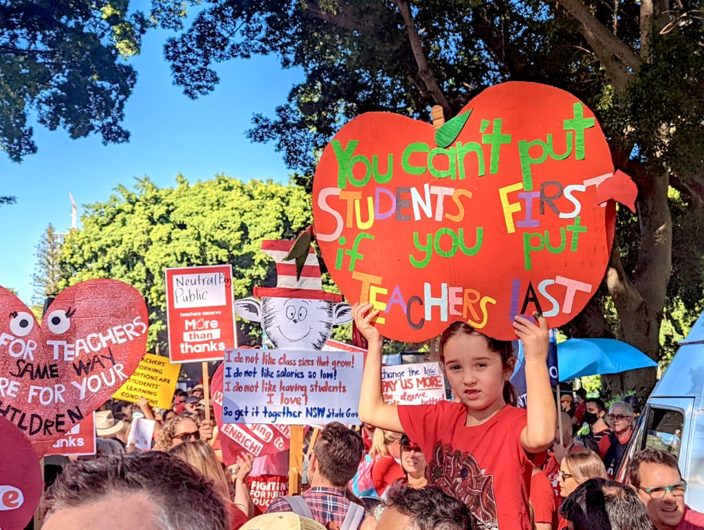 A child sits on the shoulders of a protesting teacher and holds an apple shaped sign that reads "You can't put students first if you put teachers last"