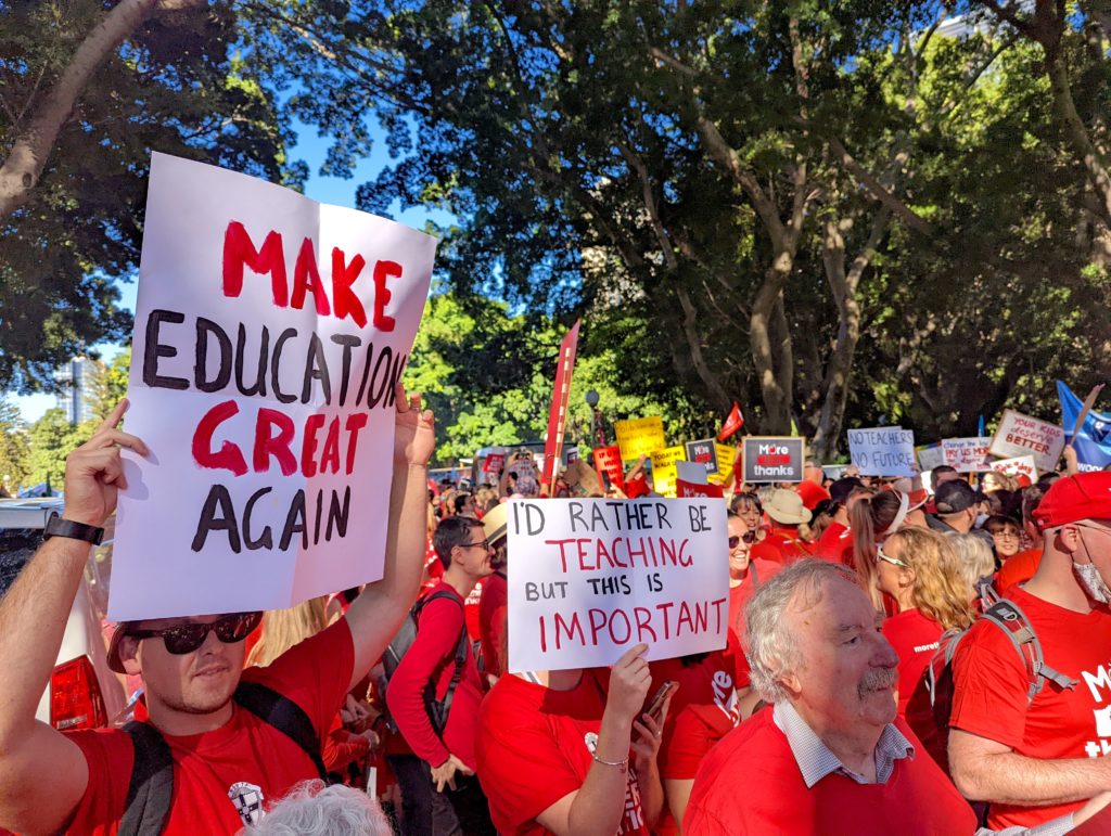 Protesting teachers holdings signs that read "Make education great again" and "I'd rather be teaching but this is important".