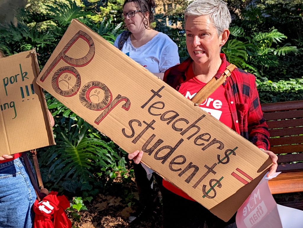 A teacher holds a sign that reads "Poor teachers = poor students"