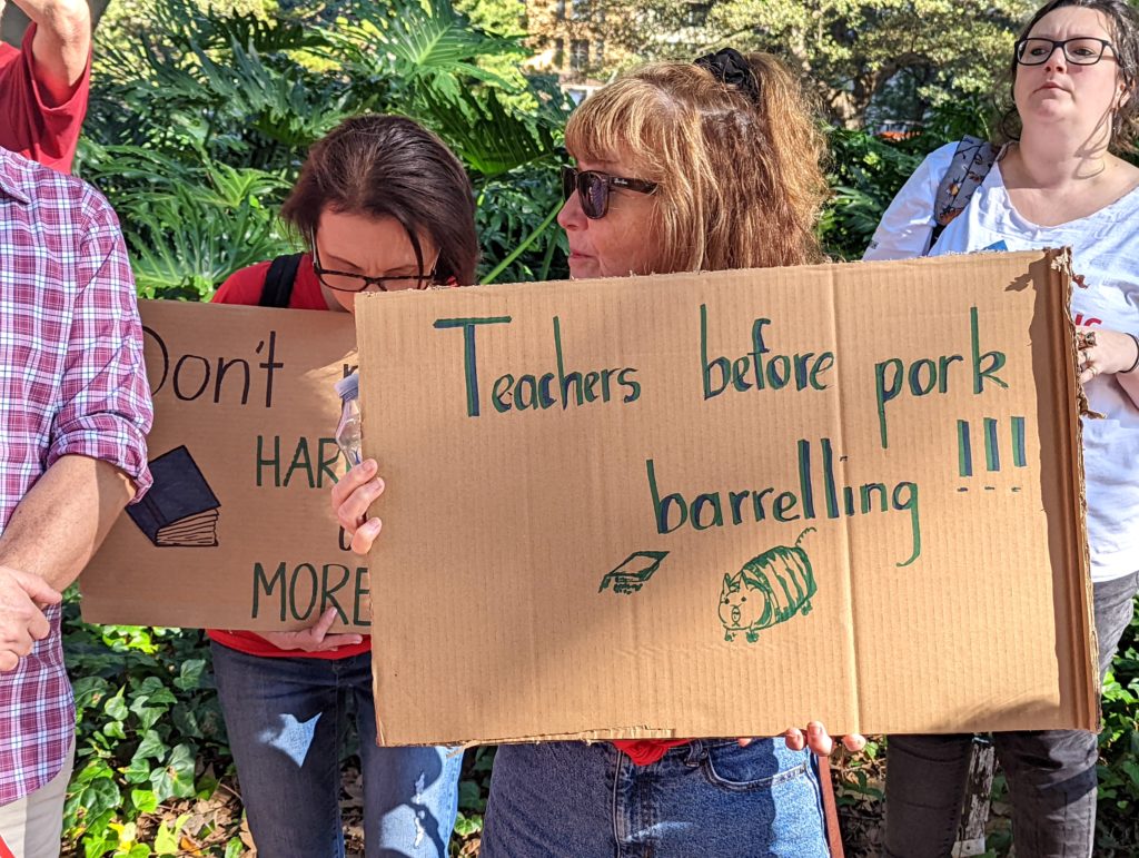 A teacher holds a sign that reads "teachers before pork barrelling"
