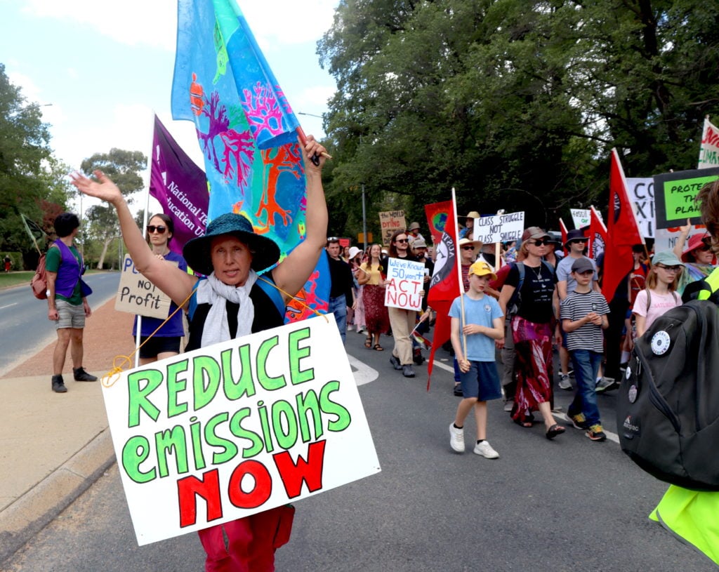 A woman walks with arm raised and wearing a sign reading 'reduce emissions now' in front of climate protesters carrying union banners