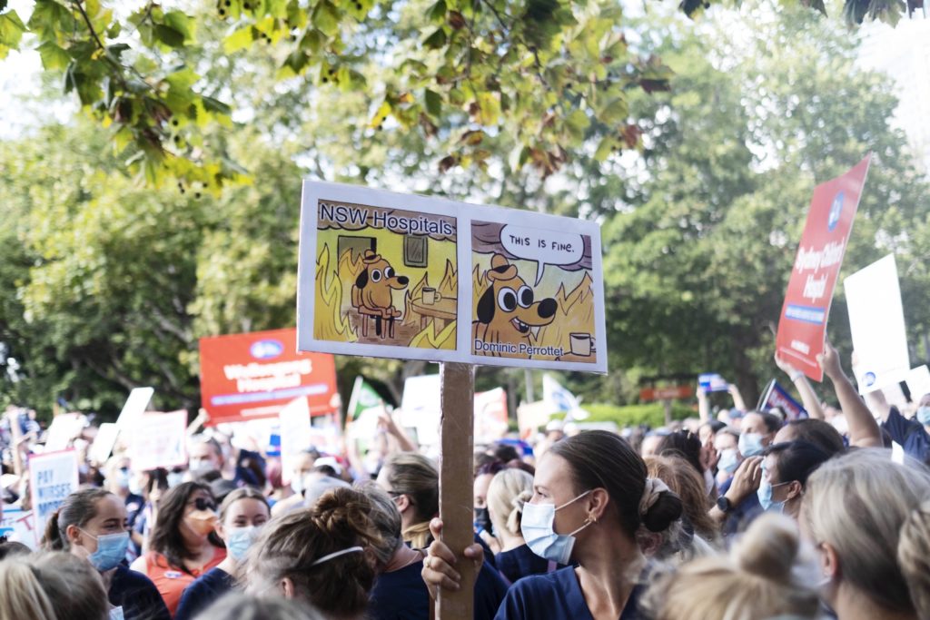 NSW nurses on strike. A nurse holds up a sign with the 'This is fine' meme symbolising Dominic Perrottet sitting doing nothing while NSW hospitals go up in flames. 