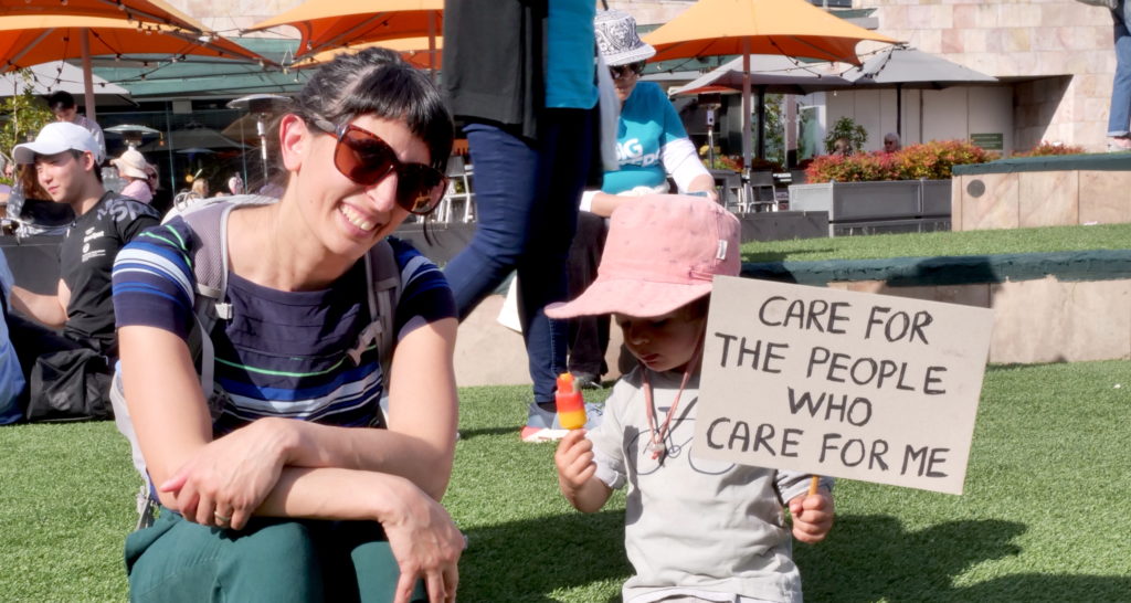 A woman sits next to a toddler who holds a sign saying, "Care for the people who care for me."