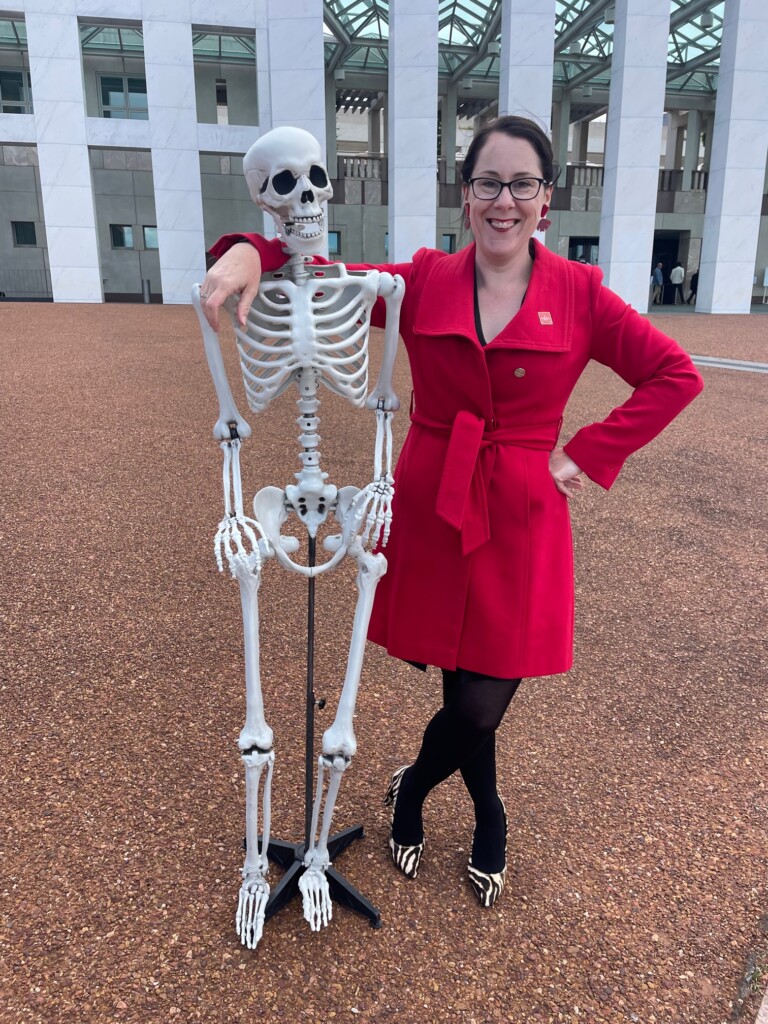 Jessica Munday stands with her arm across a model skeleton outside parliament house in Canberra.